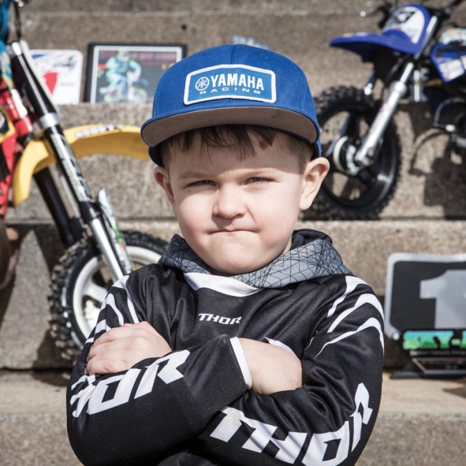 Young Boy with Arms Folded Standing In Front of Dirt Bikes